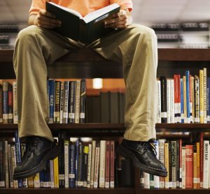 Man Reading Book and Sitting on Bookshelf in Library --- Image by © Royalty-Free/Corbis